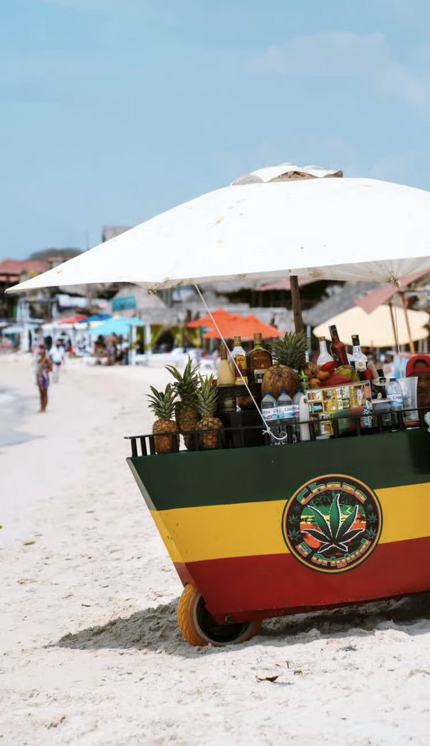A painted drinks cart parked on the sand under a white parasol, stacked with pineapples, bottles and cold drinks.