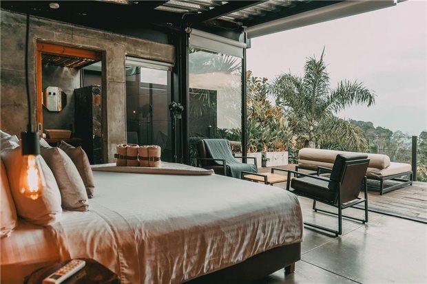 Inside a suite: bed facing a wall of glass that folds fully open onto a deck with loungers and palms.