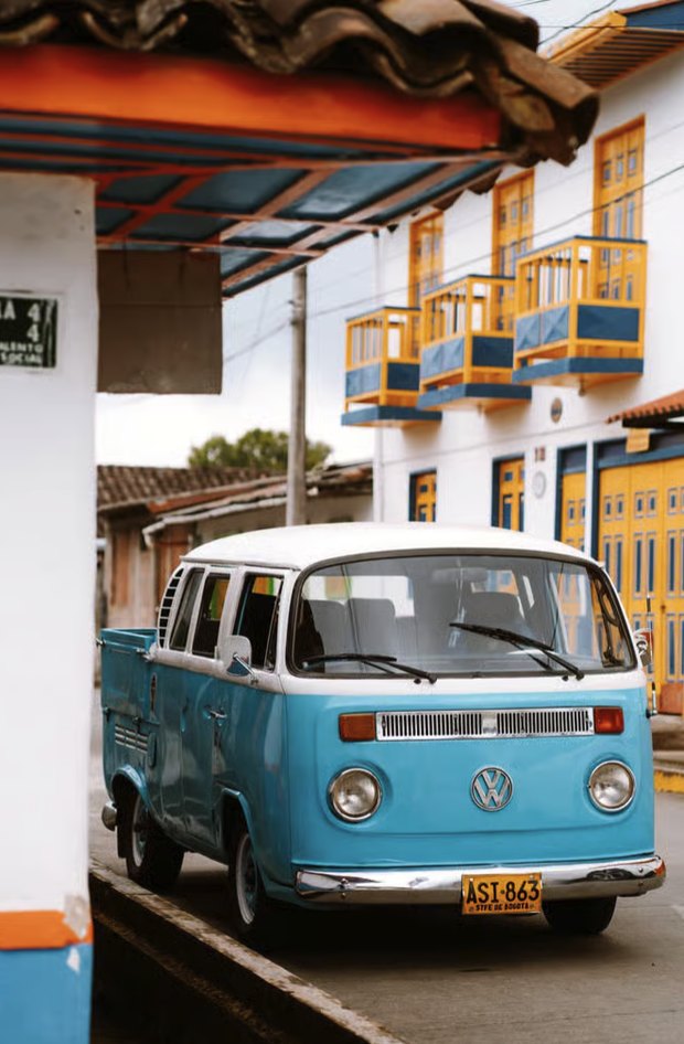 A pale blue VW camper parked on a Salento street corner, under white buildings with bright yellow and blue balconies.