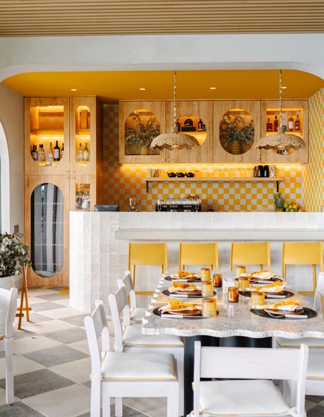 The restaurant: a marble table laid for lunch, yellow bar stools, a yellow-and-white checkerboard tiled bar and rattan pendant lamps.