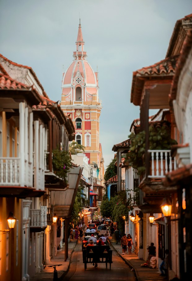 A narrow street at dusk under lit lanterns, wooden balconies either side, and the cathedral's pink dome and bell tower closing the view.
