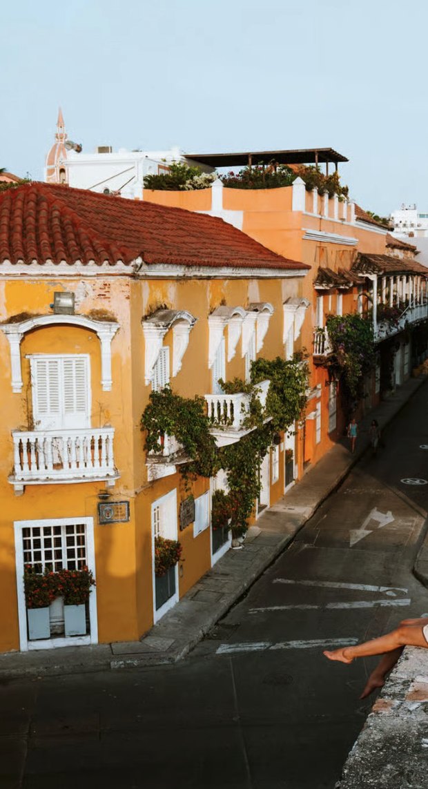 Looking down from the city walls at sunset over a yellow colonial house with white balconies and bougainvillea, a pair of bare legs dangling in the corner of the frame.