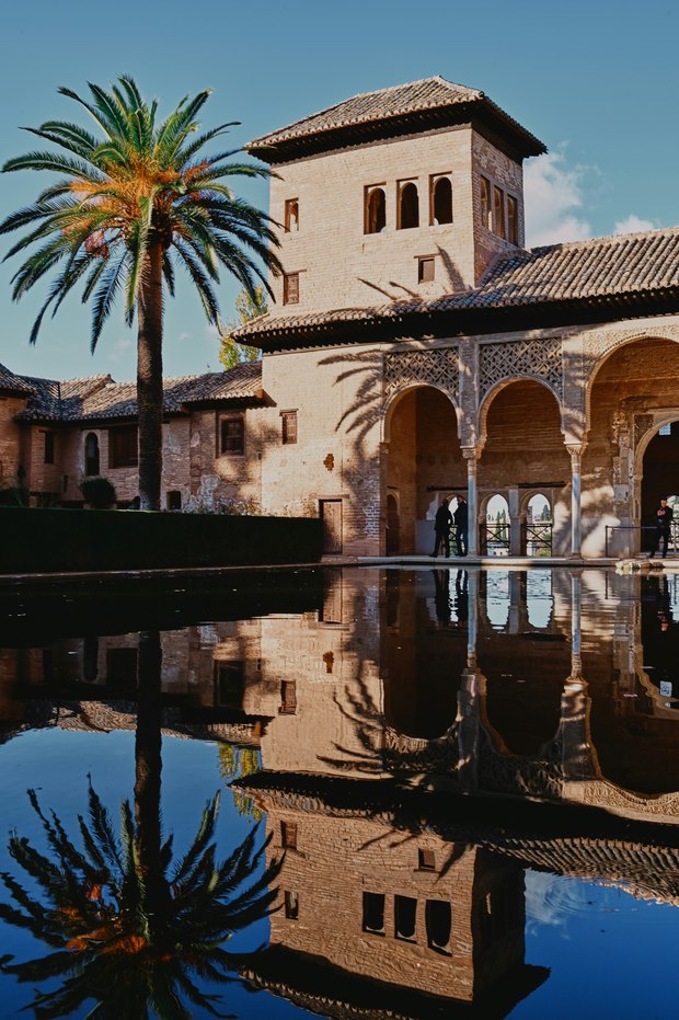 The Partal: an arcaded tower and a palm tree reflected in a still pool under a blue sky.