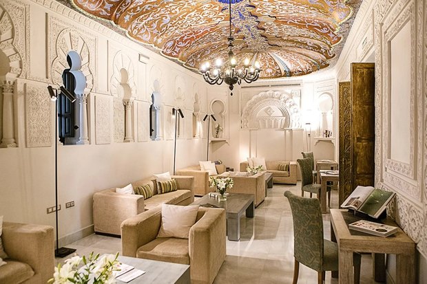A lounge with white carved Moorish arches and a gold-and-blue painted ceiling under a black chandelier.