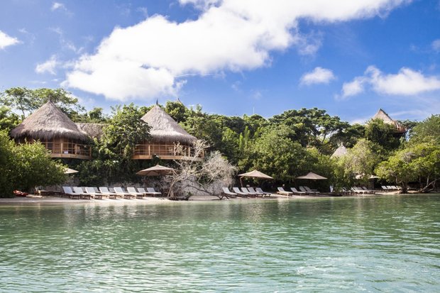Seen from the water: thatched bungalows raised among the trees above a line of sun loungers on a small beach.