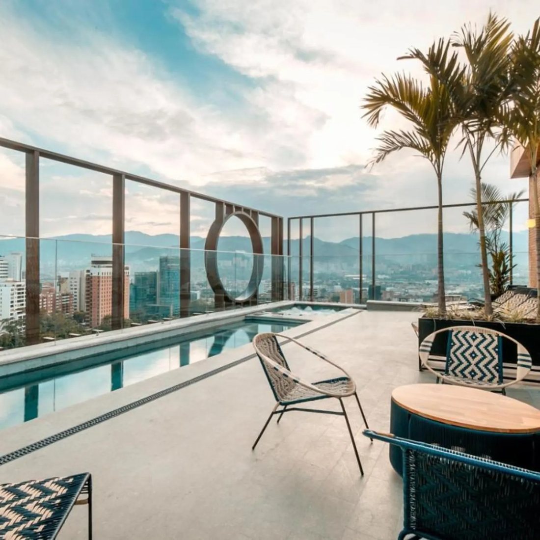 The Marquee rooftop: a long lap pool and hot tub behind glass balustrades, palms in planters, and the Medellín skyline with the Andes behind it.