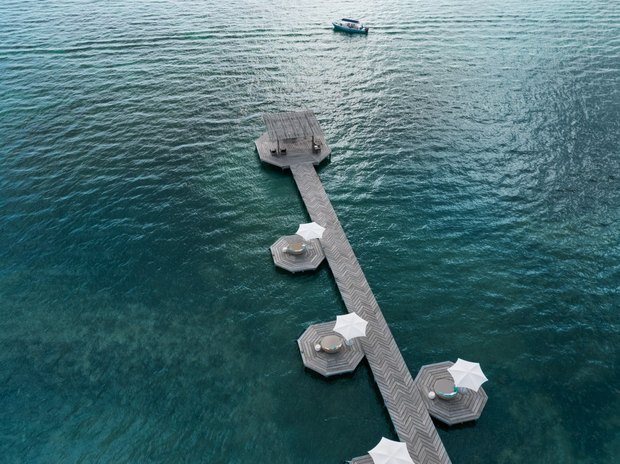 The pier from above: a herringbone boardwalk over deep green water with octagonal decks, round daybeds and white umbrellas.