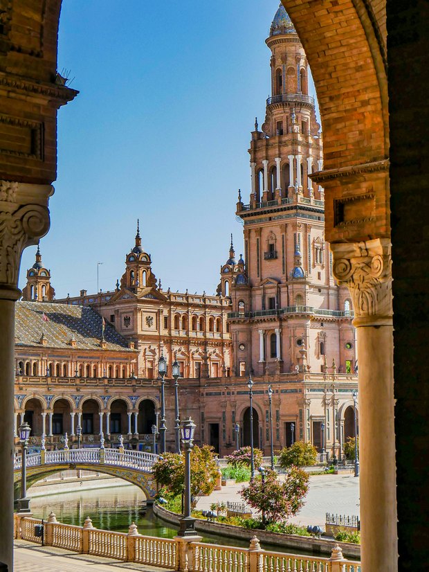 Plaza de España through a brick arch: a tall ornate tower, arcades and a tiled bridge over the canal.